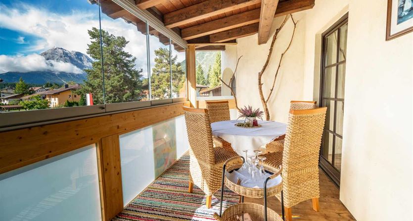 Een gezellige balkon met houten bekleding en een tafel, omringd door rotan stoelen. Op de achtergrond zijn groene bomen en bergen onder een blauwe lucht te zien.