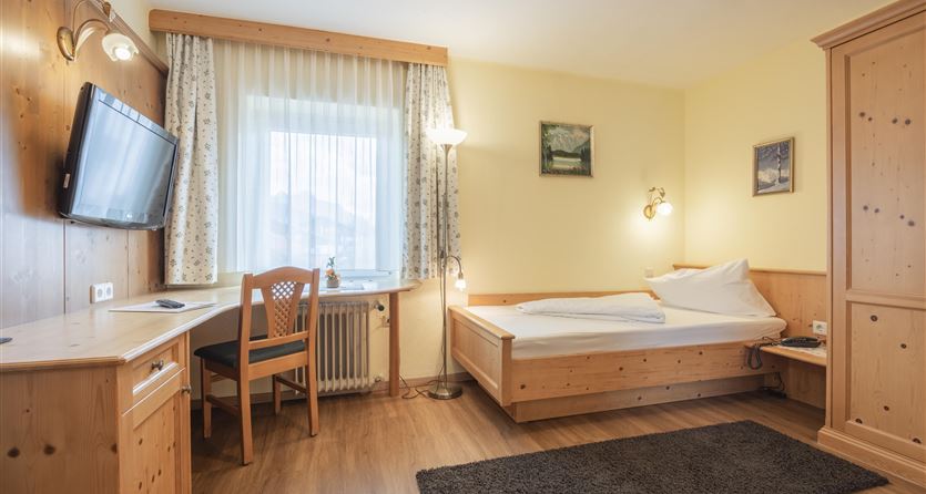 A simple room with a single bed, a desk, and a television. The walls are in warm wood tones, and the window lets in a lot of light.