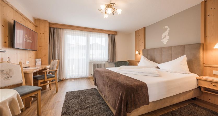 A modern hotel room with a large bed, wooden paneling, and a cozy atmosphere.  
There is a large window with a view outside and a balcony.