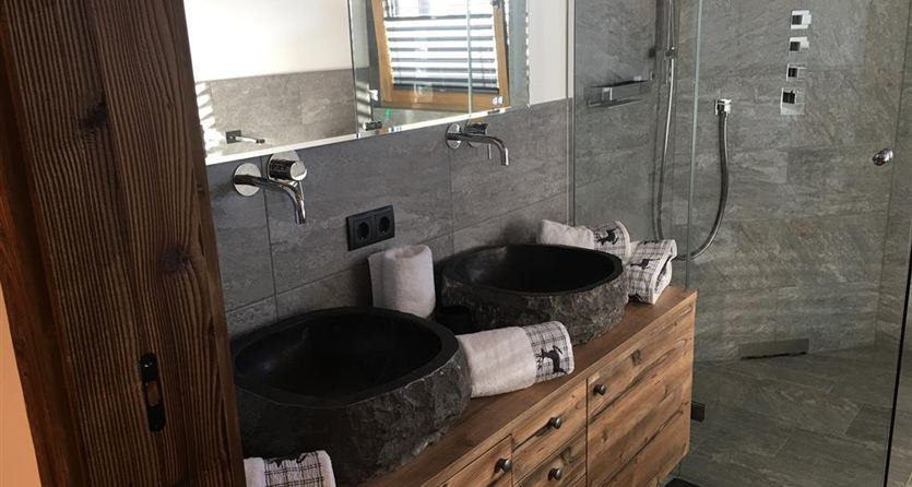 A modern bathroom with two elegant stone sinks. The shower is made of glass and the walls are designed in a gray stone look.