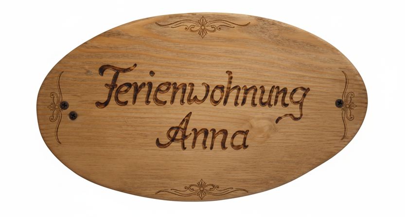 Ein hölzernes Schild mit der Aufschrift “Ferienwohnung Anna”. Es hat eine ovale Form und dekorative Details.