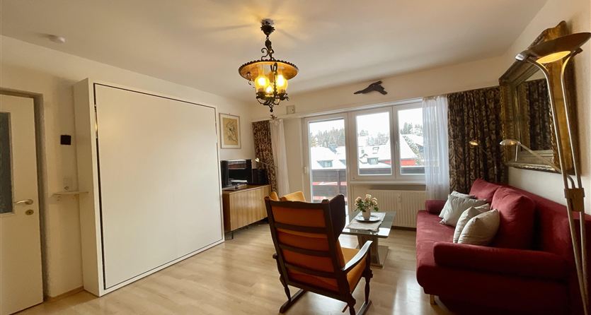 Ein gemütliches Wohnzimmer mit einem roten Sofa und einem gemütlichen Sessel. Große Fenster bringen viel Tageslicht in den Raum.