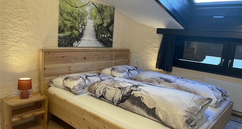 Ein gemütliches Schlafzimmer mit einem Doppelbett und holzfarbenen Möbeln. Ein großes Fenster mit einem Ausblick und ein Wandbild einer Baumallee schaffen eine einladende Atmosphäre.