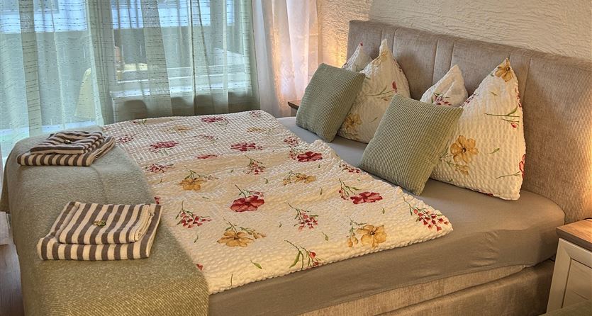 Een gezellige slaapkamer met een groot bed en decoratieve kussens. De kamer heeft grote ramen die veel daglicht binnenlaten.