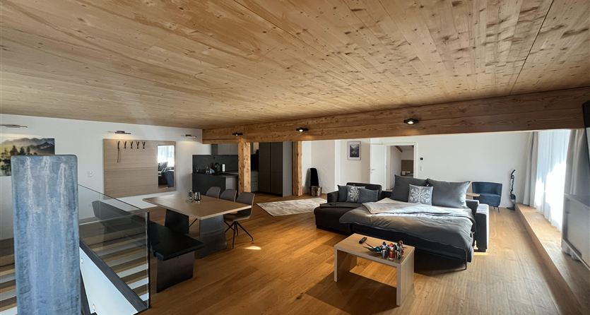 Ein modernes Wohnzimmer mit Holzdecken und einem gemütlichen Sofa. Es gibt einen klaren Blick auf den Eingang und eine offene Treppe.