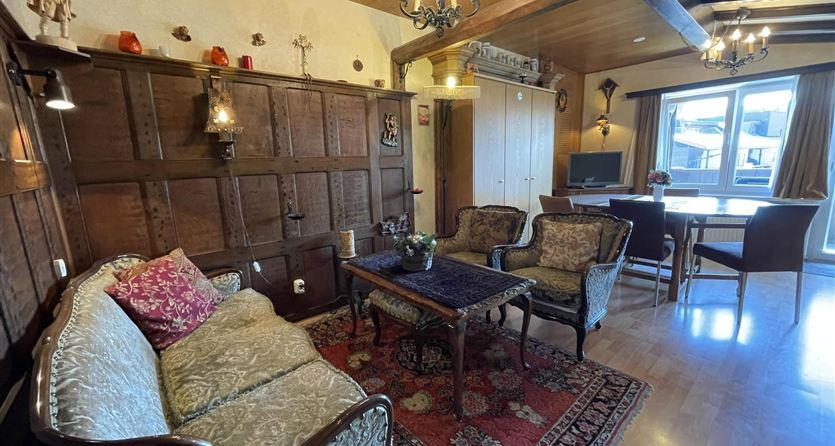 Ein gemütliches Wohnzimmer mit einem eleganten Sofa, einem Holztisch und dekorativen Stühlen. Das Zimmer ist hell mit Fenstern und traditionellem Holzdekor.