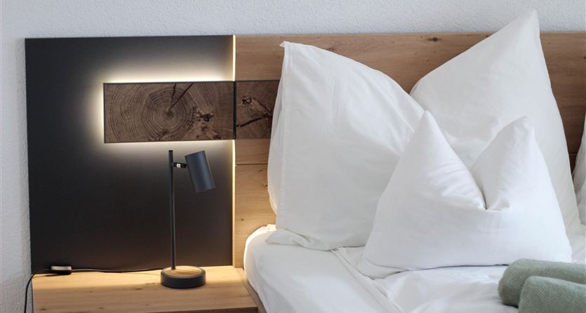A stylish bedroom with a bed, white pillows, and a modern lamp on a nightstand. The wall behind the headboard is designed with wood and an elegant light source.