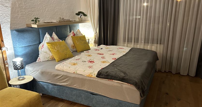 Een gezellig slaapkamer met een groot bed, kleurrijke kussens en een zachte dekbed. Het bed staat voor een raam met gordijnen en een moderne lamp.