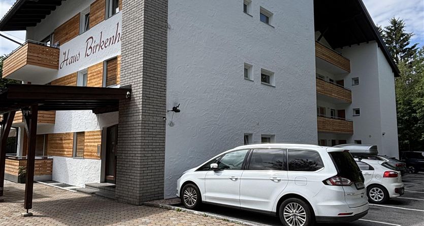 Un edificio moderno con diversi piani e rivestimento in legno. Davanti alla casa ci sono due auto su un parcheggio pavimentato.