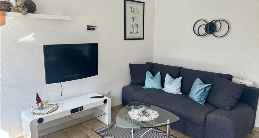 Un soggiorno moderno con un divano grigio e cuscini colorati.  
Un televisore a schermo piatto e un tavolo rotondo in vetro completano l'arredamento accogliente.