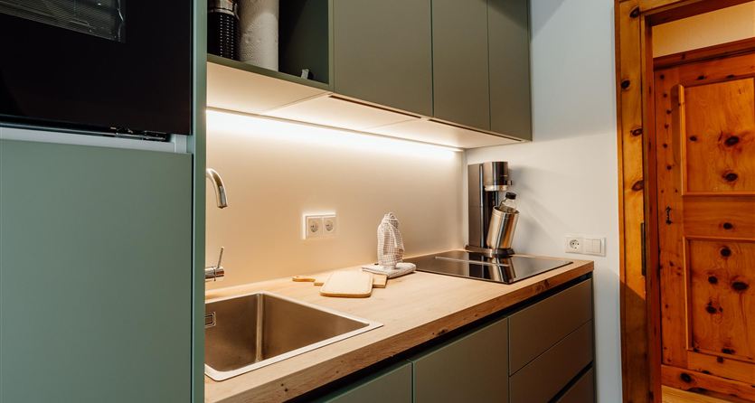 Eine moderne Küche mit grünen Schränken und einer Holzarbeitsplatte. Die Ausstattung umfasst ein Spülbecken und Kochfeld, umgeben von warmer Beleuchtung.