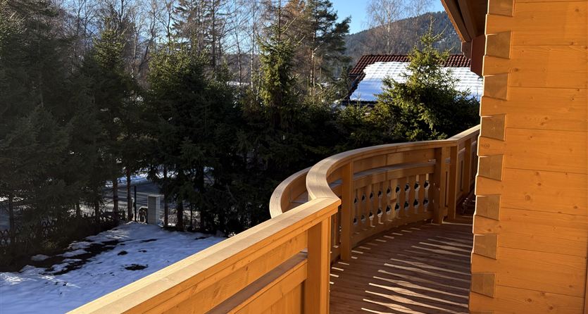 Een houten balkon met een gebogen leuning. Op de achtergrond zijn bomen en een blauwe lucht te zien.