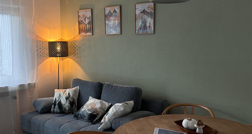 A cozy living space with a blue sofa and a round wooden table. Pictures hang on the wall and a lamp provides pleasant light.