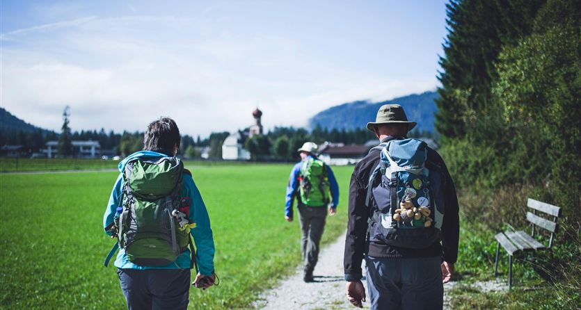 Drie wandelaars met backpacks lopen op een pad door groene weilanden. Op de achtergrond zijn bomen en bergen te zien.