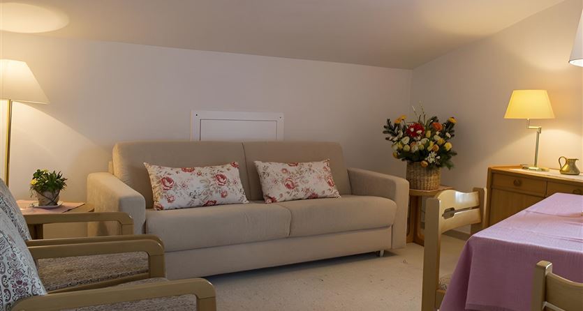 Ein gemütliches Wohnzimmer mit einem beigen Sofa und dekorativen Kissen. Es gibt einen Tisch und einige Pflanzen sowie weiche Beleuchtung.