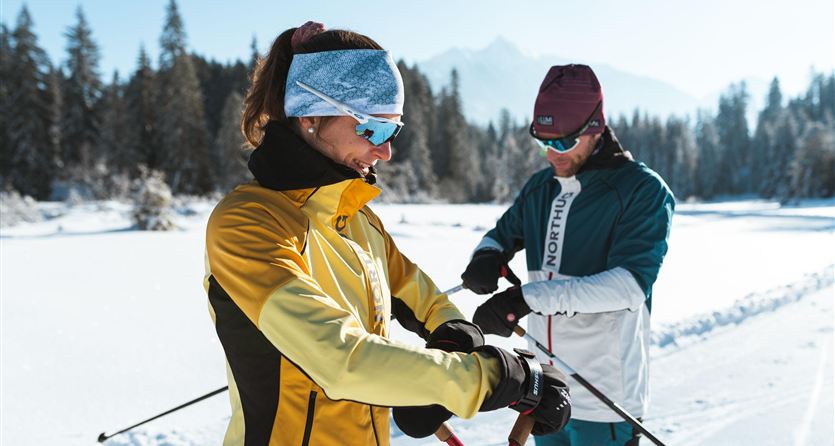 Ein Paar in winterlicher Sportkleidung präpariert ihre Skiausrüstung im Schnee. Der klare Himmel und die schneebedeckten Bäume im Hintergrund schaffen eine schöne Winterlandschaft.