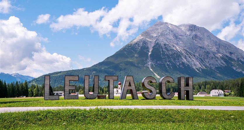 A sign with the inscription "LEUTASCH" stands in front of a large mountain. The surroundings are green with meadows and a blue sky.