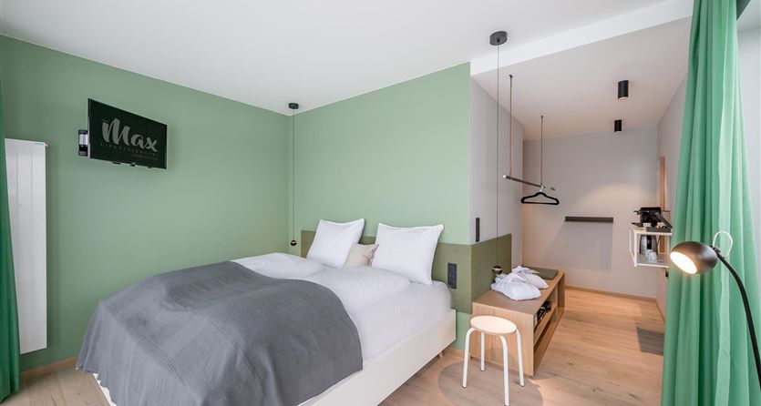 Ein modernes Schlafzimmer mit grünen Wänden und warmem Holzfußboden. Das Zimmer ist minimalistisch eingerichtet mit einem Doppelbett, einem Hocker und einem Fernseher.