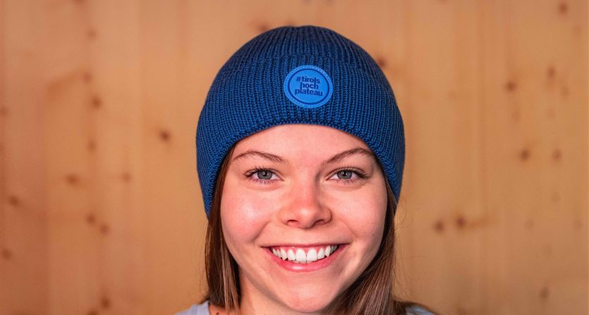 Eine lächelnde Frau trägt eine blauen Mütze. Im Hintergrund ist eine Holzwand sichtbar.