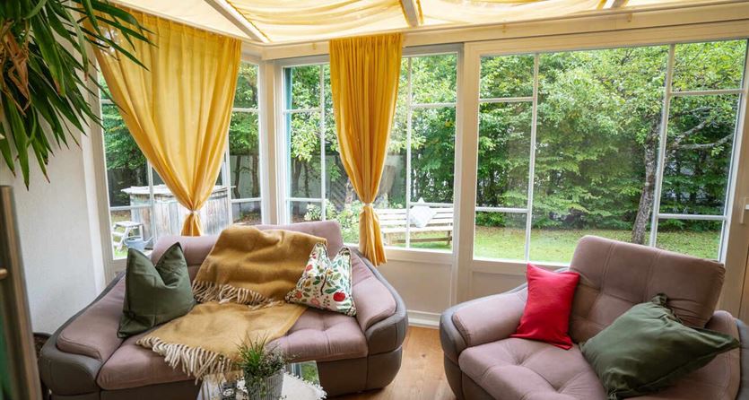 Een gezellige woonkamer met grote ramen en gele gordijnen. Er zijn comfortabele banken en uitzicht op de tuin.
