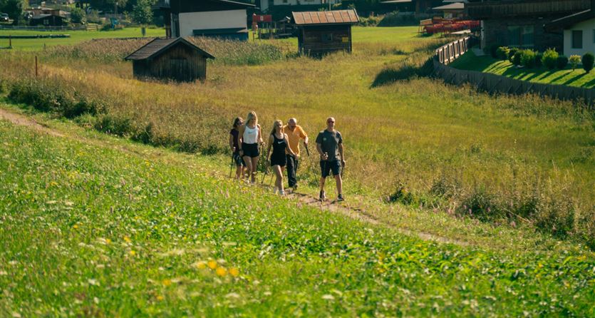 Eine Gruppe von fünf Personen wandert auf einem grünen Feld. Im Hintergrund sind traditionelle Häuser und ein blauer Himmel sichtbar.