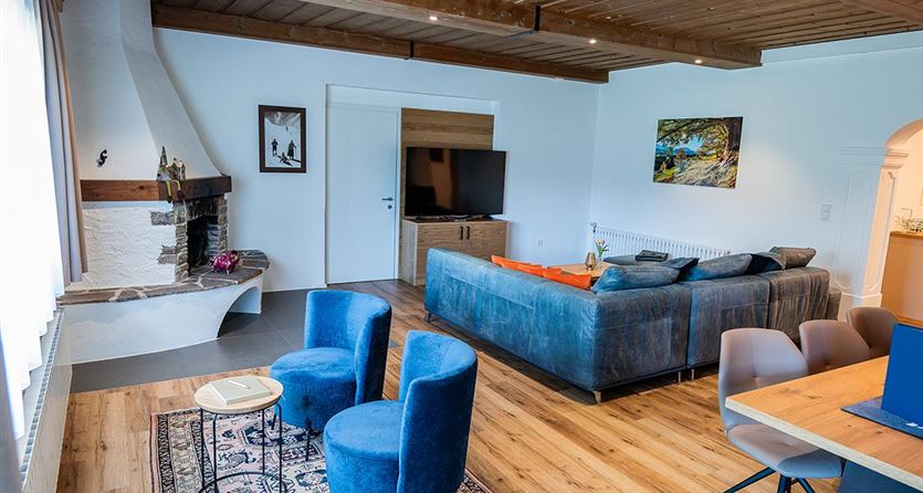 Een moderne woonkamer met houten plafond en een gezellige inrichting. Er zijn blauwe stoelen, een bank en een eettafel in een lichte ruimte.