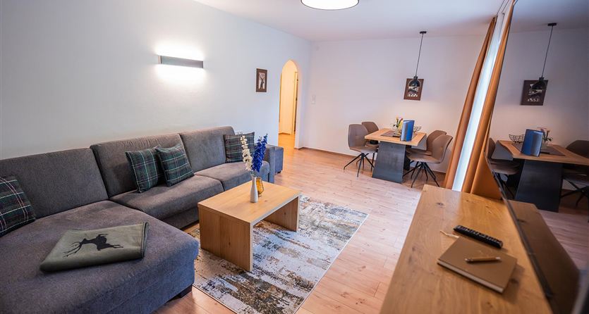 Een gezellige woonkamer met een grijze bank en een houten tafel. De ruimte is licht en modern ingericht, met een kleine eethoek op de achtergrond.