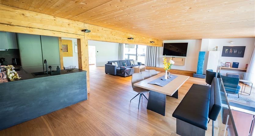 Een moderne, stijlvolle woonkamer met houten vloer en grote ramen. De inrichting omvat een bank, een eettafel en een flatscreen-tv.