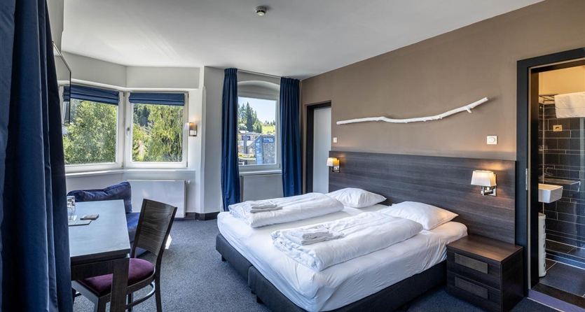 A modern hotel room with two single beds, a table, and a chair.  
Large windows ensure plenty of light and a view outside.