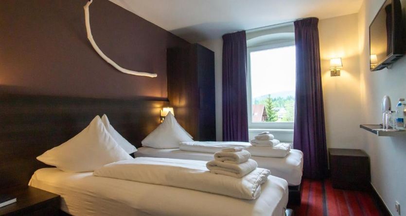 A modern room with two single beds and white bedspreads. The room has a window with a view and a television on the wall.