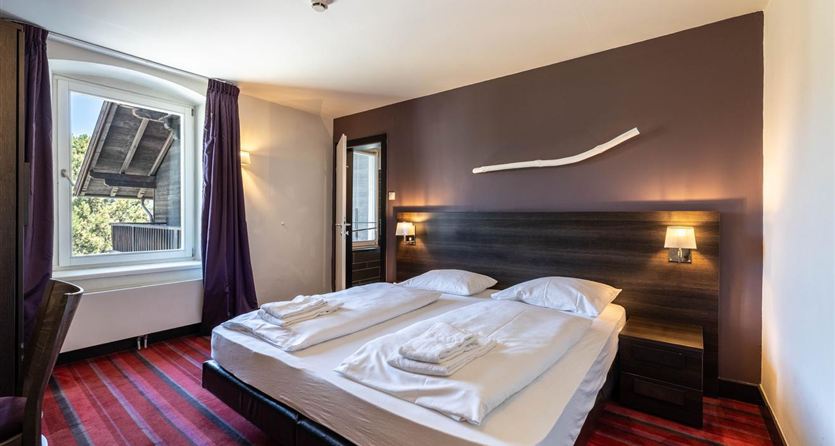A modern hotel room with two single beds and stylish lamps. The room has windows with a view and dark curtains.