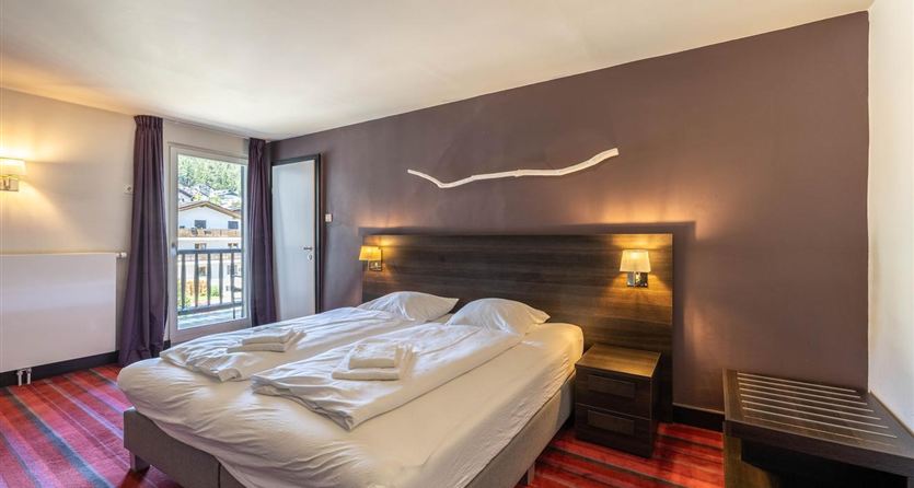 A cozy hotel room with a double bed and modern furnishings. Large windows provide plenty of daylight and a view outside.