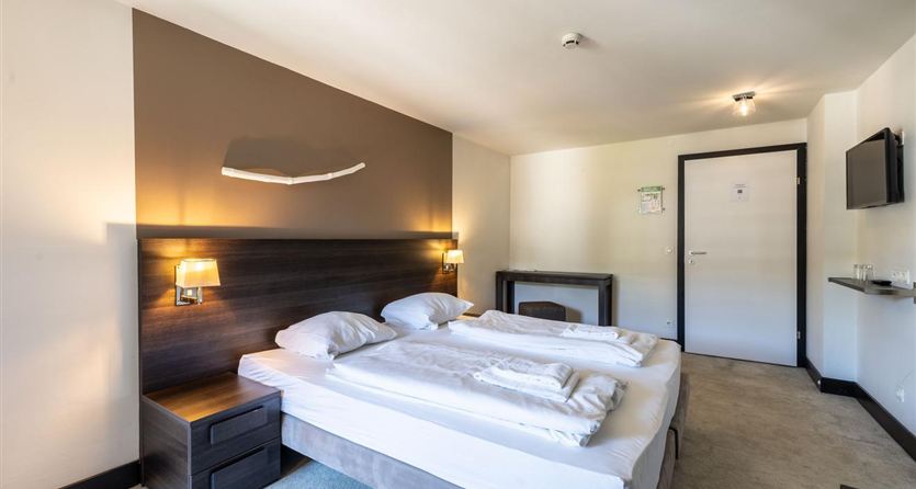 A modern bedroom with two beds and bright furnishings. On the wall, there are bedside lamps and a television installed.