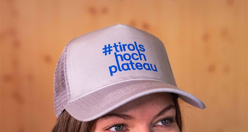 Eine Person trägt eine graue Kappe mit dem Aufdruck "#tirols hochplateau" in blauer Schrift. Der Hintergrund ist unscharf und hat eine natürliche Holzoptik.