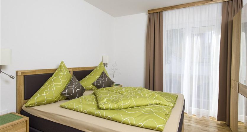 Een moderne slaapkamer met een tweepersoonsbed en groene beddengoed. Heldere gordijnen laten daglicht binnen en zorgen voor een uitnodigende sfeer.