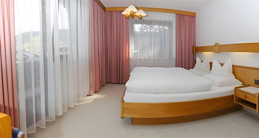 Ein gemütliches Schlafzimmer mit einem Doppelbett und hellen Vorhängen. Das Zimmer hat einen warmen Holzlook und viel natürliches Licht.