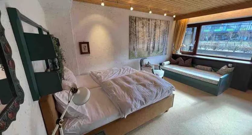 Ein gemütliches Schlafzimmer mit einem Doppelbett und einer Leseecke. Große Fenster sorgen für viel natürliches Licht und Blick auf die Umgebung.