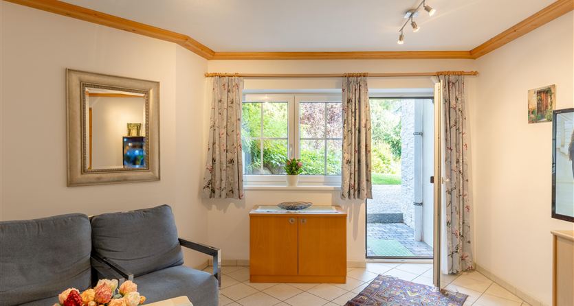 Eine helle und einladende Wohnraumecke mit einem grauen Sofa und einem Sideboard. Durch das Fenster sieht man einen grünen Garten.