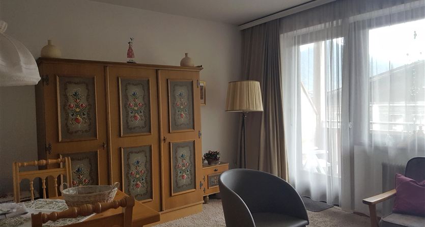 Ein gemütlicher Wohnraum mit einem klassischen Kasten und einer Stehlampe. Große Fenster sorgen für viel Tageslicht.