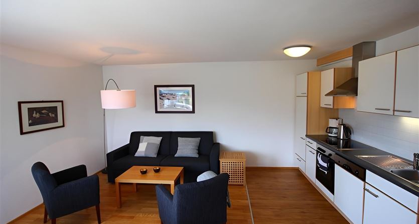 Ein helles Wohnzimmer mit einem schwarzen Sofa und zwei blauen Stühlen. Die Küche ist modern und gut ausgestattet, mit einem Tisch in der Mitte des Raumes.