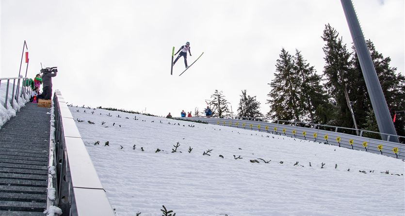 Un saltatore con gli sci salta da un trampolino coperto di neve. Sullo sfondo si vedono spettatori e alberi.