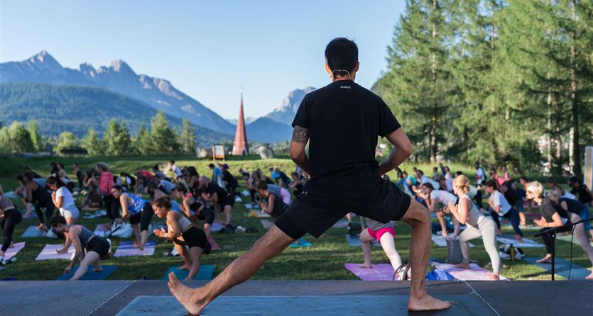 Un evento di yoga all'aperto con molti partecipanti. Le montagne sullo sfondo e gli alberi creano un'atmosfera tranquilla.