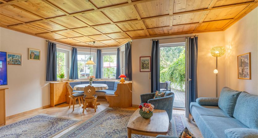 Ein gemütliches Wohnzimmer mit blauen Sofas und einem Holztisch. Große Fenster lassen viel Licht herein und bieten einen Blick auf den Garten.