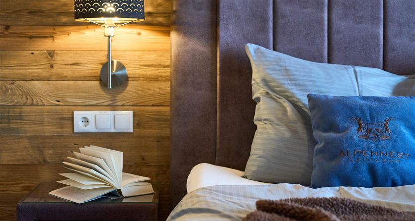 A cozy bedroom with a large bed and pillows. On the bedside table lies an open book and a lamp provides warm light.