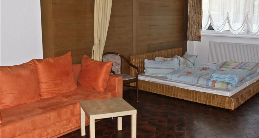 Una stanza semplice con un divano arancione, un tavolo e un letto. Le pareti sono rivestite di legno e ci sono grandi finestre con tende.