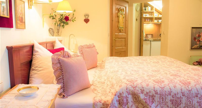 A cozy bedroom with a large bed and colorful pillows. The room is warmly lit and there is an adjacent area with a kitchen.