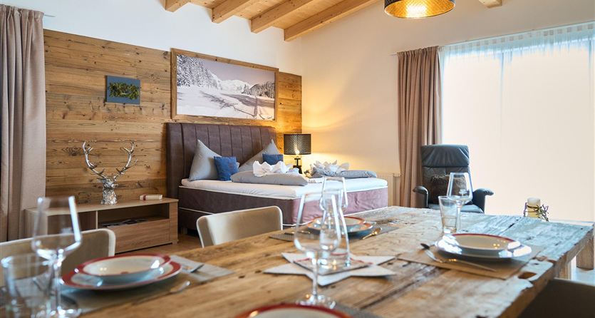 A modern room with a large bed and wooden details. At the table, there are set places with dishes and glasses.
