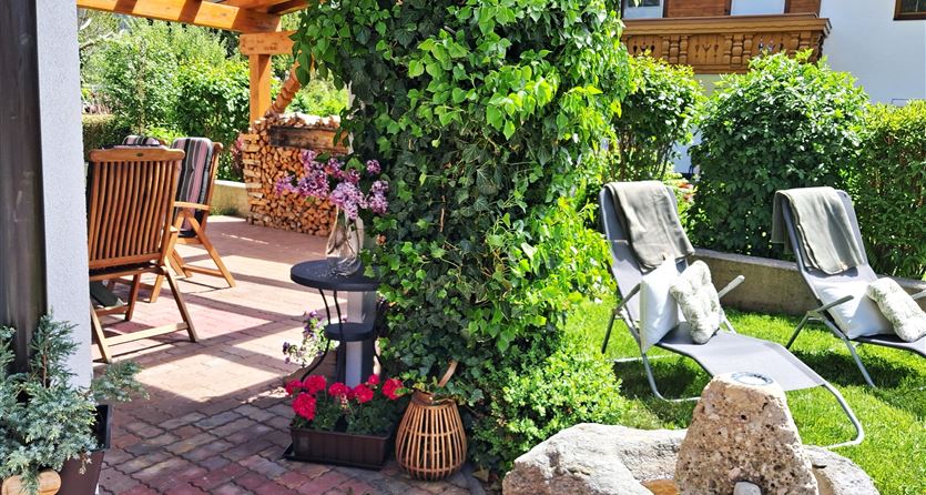 Eine gemütliche Terrasse mit Holzdecken und Gartenstühlen. Im Vordergrund steht eine grüne Pflanze und ein kleiner Steingrill.