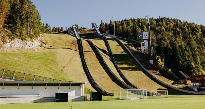 Una pista di salto con gli sci circondata da prati verdi e alberi. In primo piano si vede un campo da calcio.