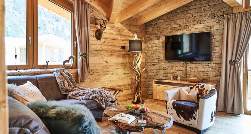 Ein gemütliches Wohnzimmer mit Holzdecke und großen Fenstern. Die Einrichtung kombiniert moderne und rustikale Elemente, darunter ein bequemer Sessel und eine elegante Couch.